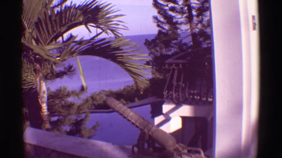 1971 Hawaii home-movie frame looking from a hotel room or balcony past palm fronds and railing toward ocean water.