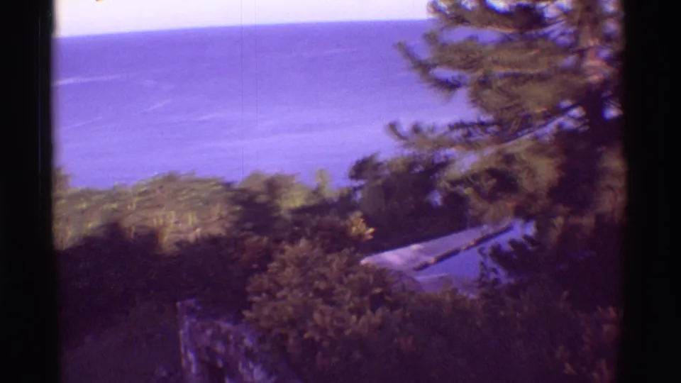 1971 Mauna Ulu, Hawaii frame showing ocean, tropical trees, and a partial roof or paved edge below the overlook.