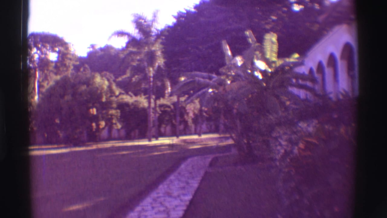 1971 Hawaii 8mm frame of a curved garden path, dense tropical plants and a white building with arched openings.