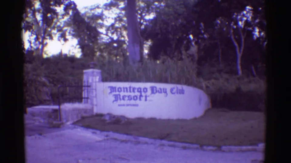 1971 8mm Hawaii travel frame showing the Montego Bay Club Resort entrance sign, gate, stone post, trees, and roadside verge.