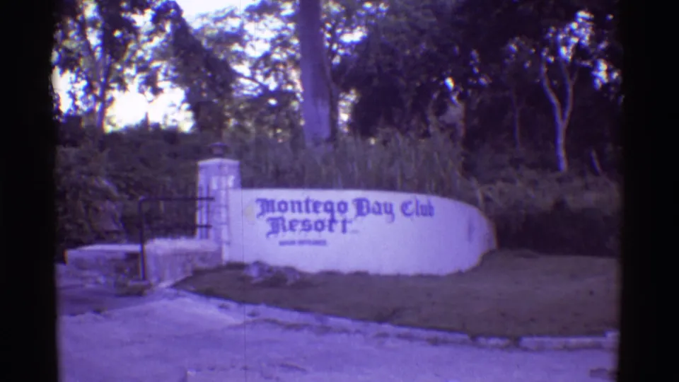 1971 Hawaii 8mm frame showing the Montego Bay Club Resort entrance sign on a white curved wall near a gate and shaded trees.