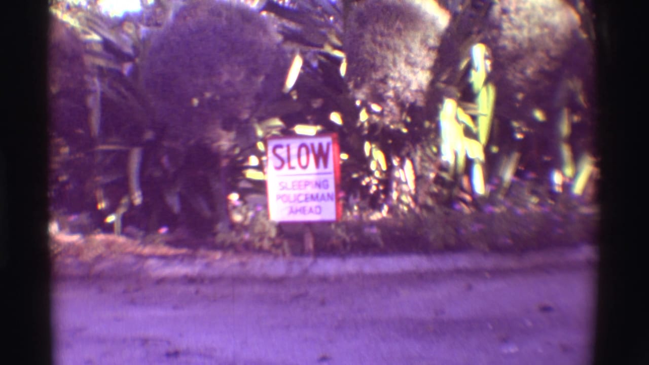 1971 Hawaii 8mm frame showing a SLOW SLEEPING POLICEMAN AHEAD sign beside a curb and lush roadside foliage.