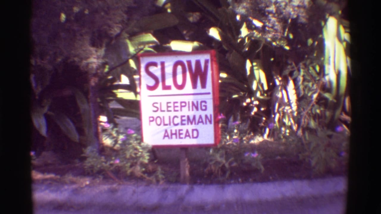 Close 1971 Hawaii 8mm view of the SLOW SLEEPING POLICEMAN AHEAD sign with tropical leaves and shrubs behind it.