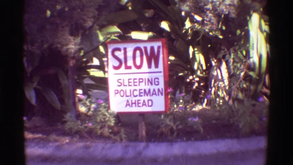 Close 1971 Hawaii 8mm view of the SLOW SLEEPING POLICEMAN AHEAD sign with tropical leaves and shrubs behind it.