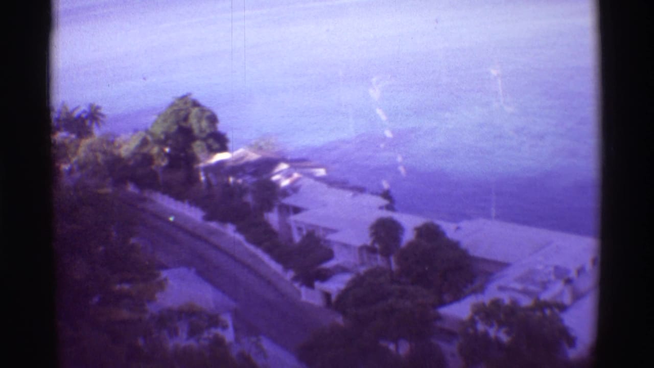 1971 Hawaii home-movie frame looking down on coastal roofs and vegetation with the ocean beyond.