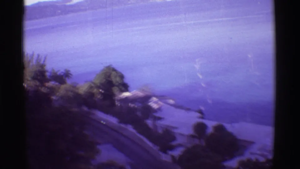 1971 Hawaii 8mm frame showing blue water, distant hills, trees, shoreline structures, and a road below the camera position.
