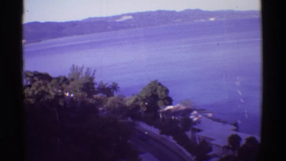 1971 Hawaii home-movie frame with tropical trees in the foreground, blue water below, and low hills across the water.