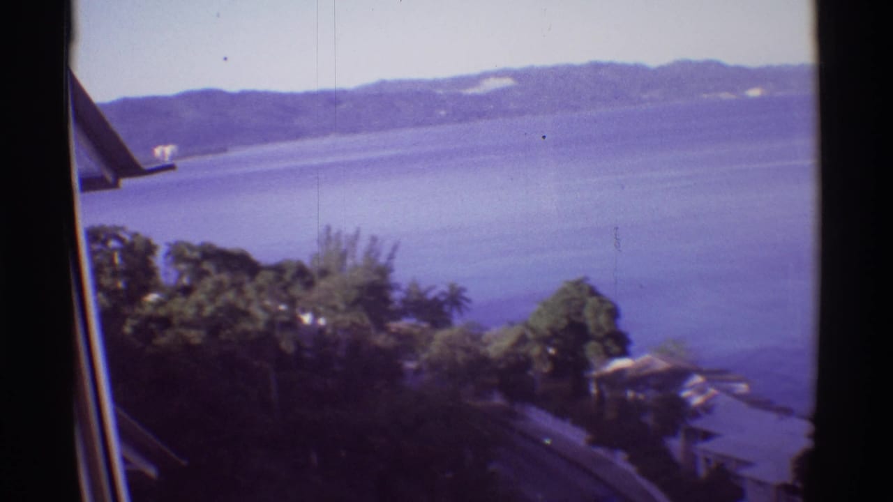 1971 Hawaii 8mm view from near a building edge, looking over vegetation, a road, shoreline structures, water, and hills.