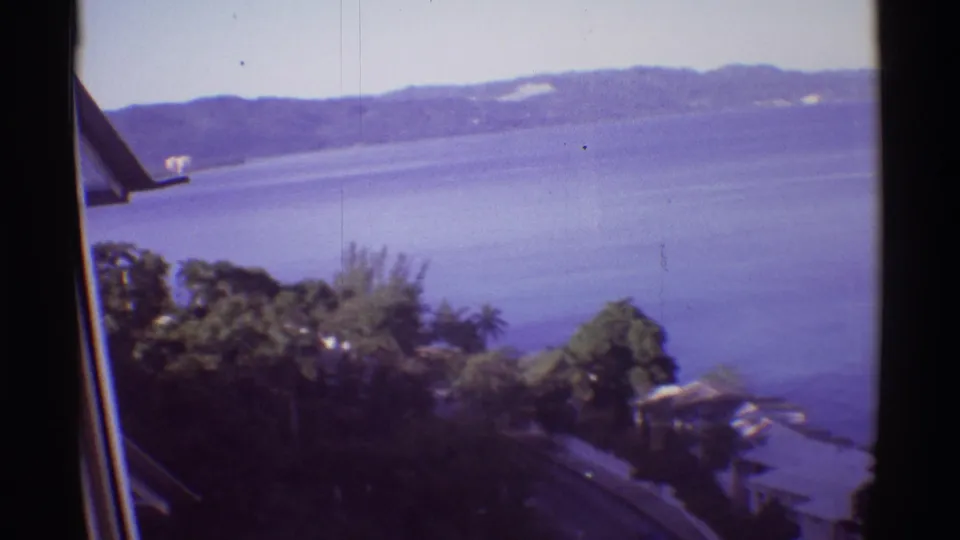 1971 Hawaii 8mm view from near a building edge, looking over vegetation, a road, shoreline structures, water, and hills.