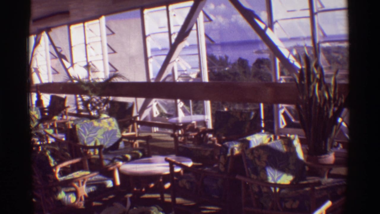 1971 Hawaii frame inside a glass-walled solarium with tropical-print seating, potted plants, and blue water visible through the windows.