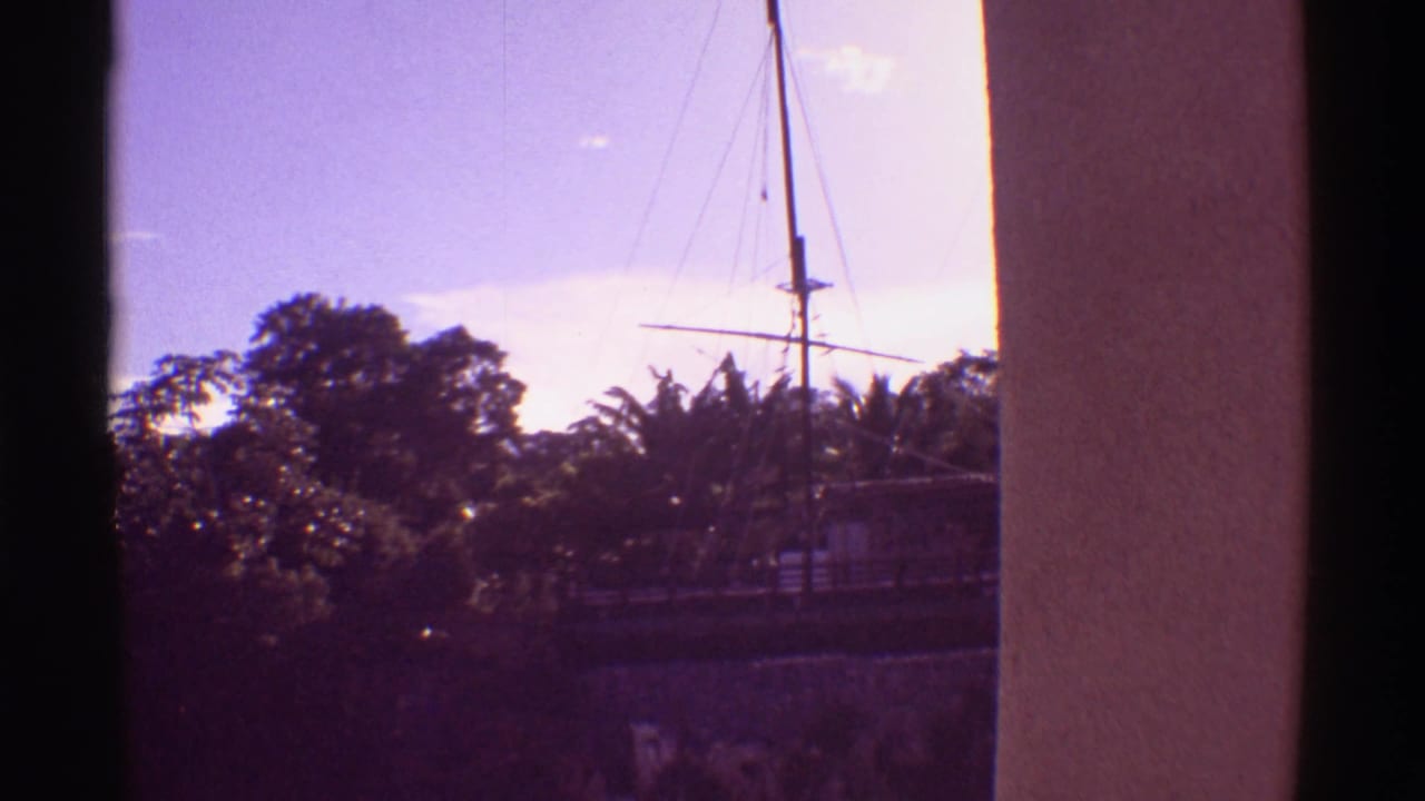 Sailboat mast, rigging, trees, and blue sky seen between vertical building edges.