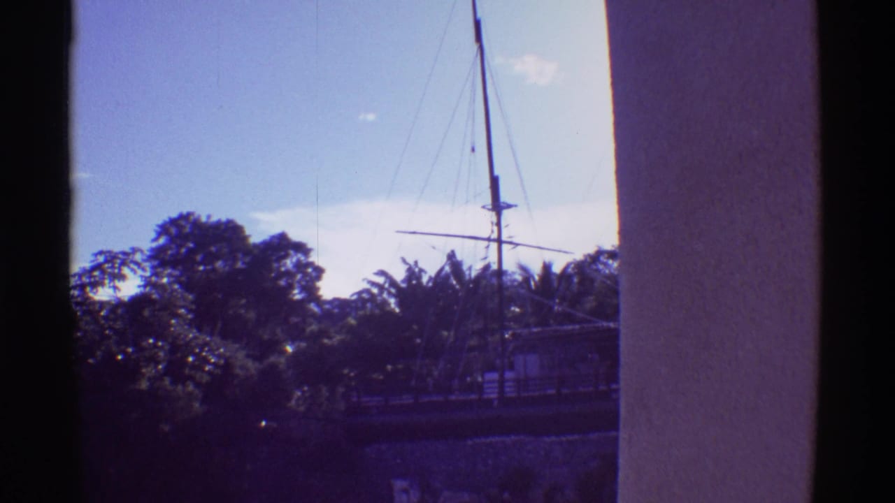 1971 Hawaii home-movie frame of a docked sailboat's mast and rigging viewed past tropical foliage and a shaded building edge.