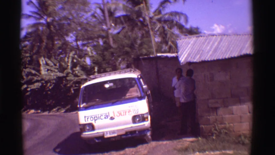 1971 Hawaii 8mm frame showing a white branded tour van near a cinder block building under palm trees.