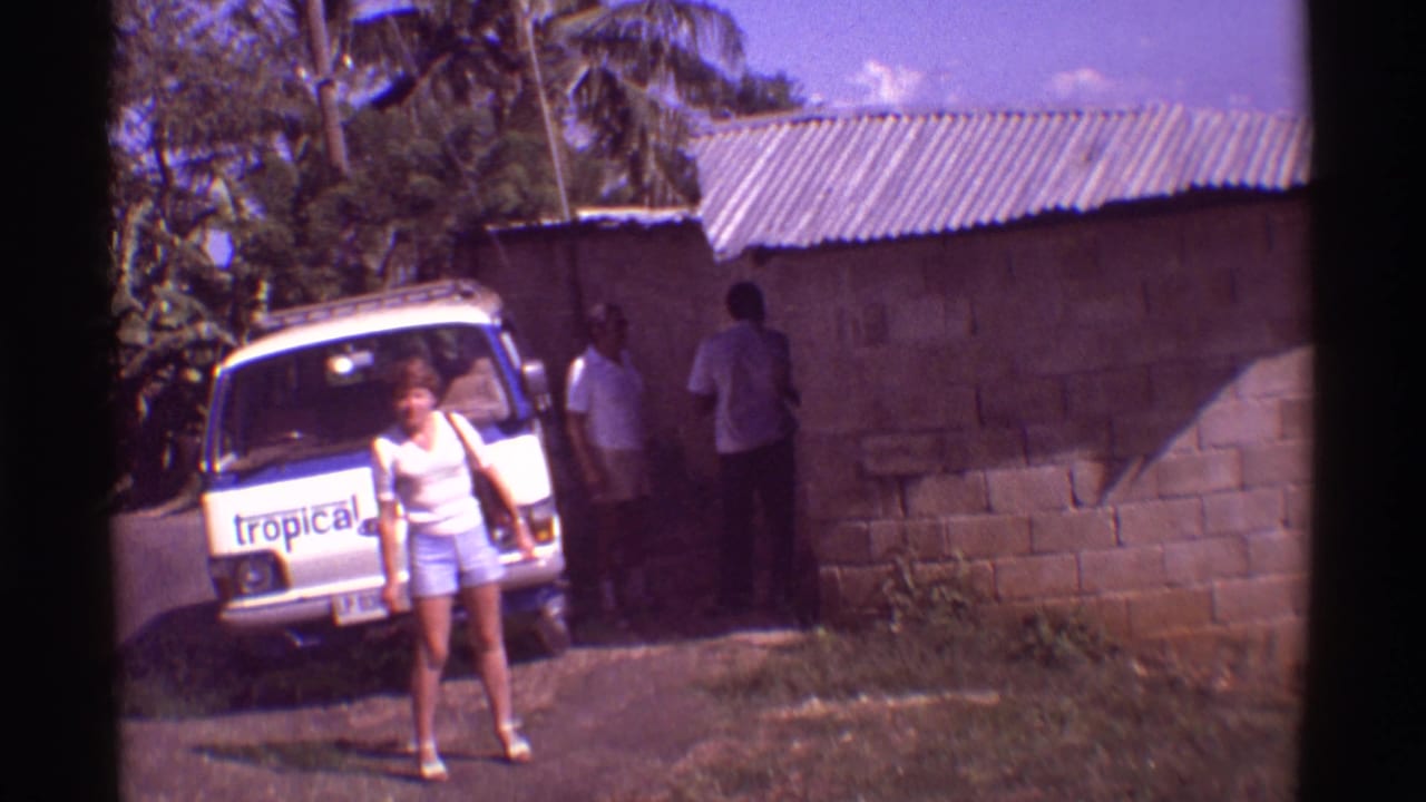 1971 Hawaii 8mm frame of visitors near a branded tour van beside a cinder block roadside building.