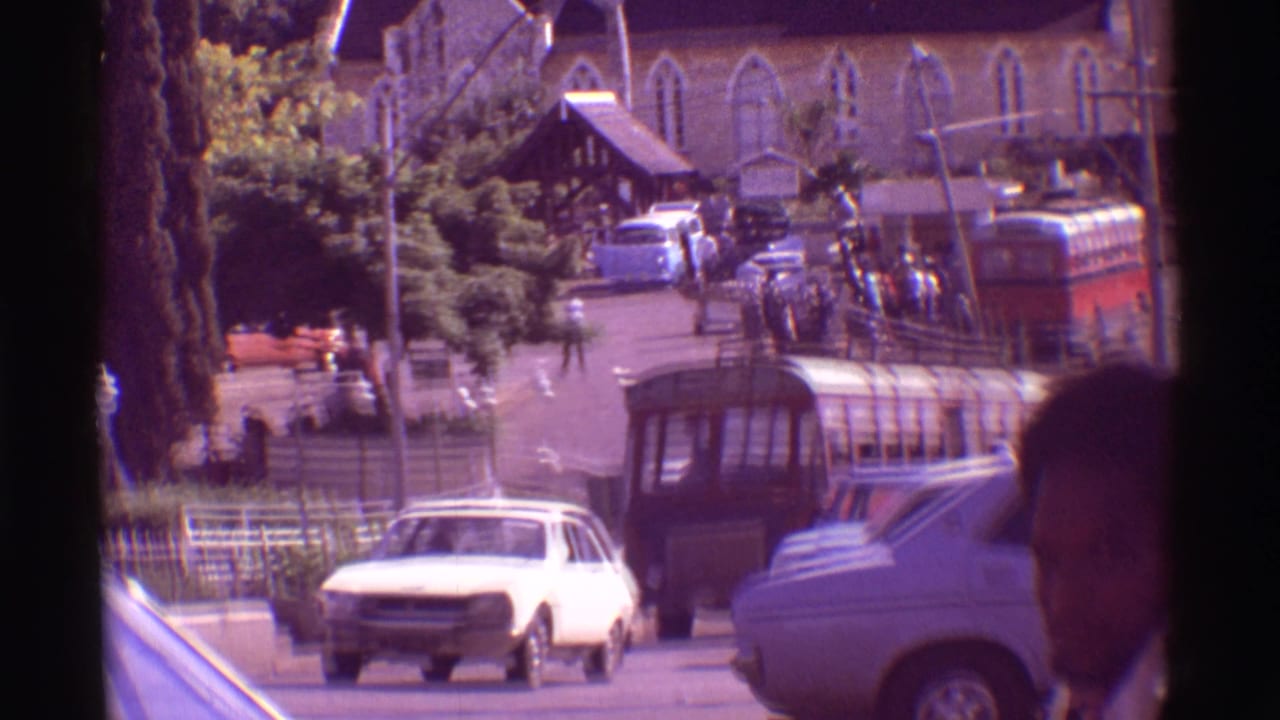 1971 Hawaii street view with vintage cars, a red bus, pedestrians, and a church-like stone building in the background; a partial face appears at right.