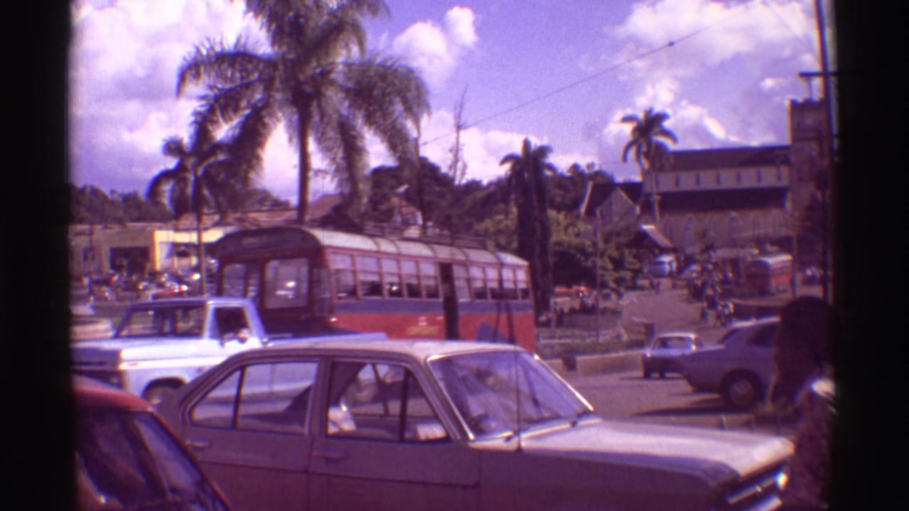 Street with palm trees, classic cars, a red-and-white bus, and pedestrians near a tropical town center.