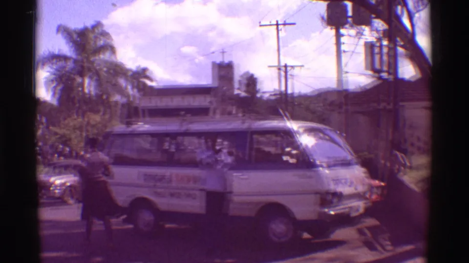 White tour van with visible branding in a 1971 Hawaii street scene, with pedestrians, palms, utility poles, and low buildings around it.