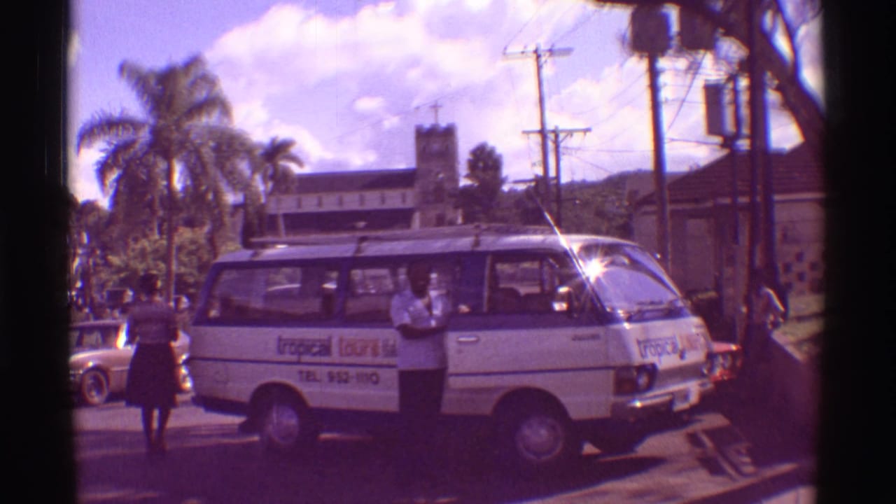 1971 Hawaii street frame showing a white tour van, a man by its side door, a woman walking past, palms, cars, and a tower-like building.