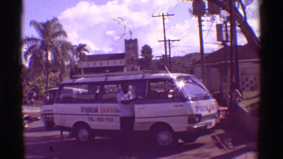 White branded tour van in a 1971 Hawaii street scene with a man at the side door, palm trees, utility poles, cars, and bright sun flare.