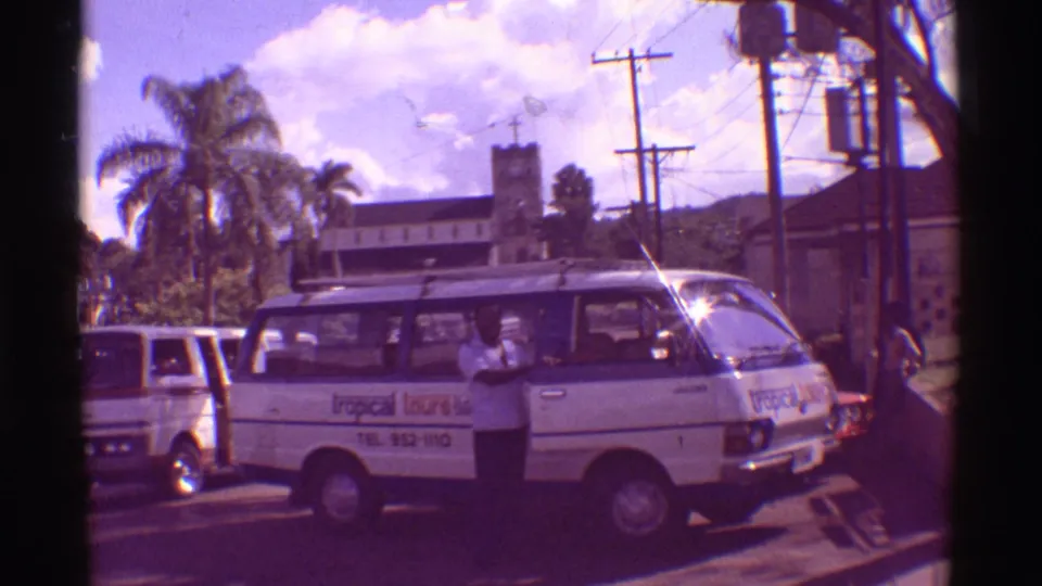 1971 Hawaii 8mm frame of a white Tropical Tours van at a street stop with a man by the open door, palms, cars, and utility poles nearby.