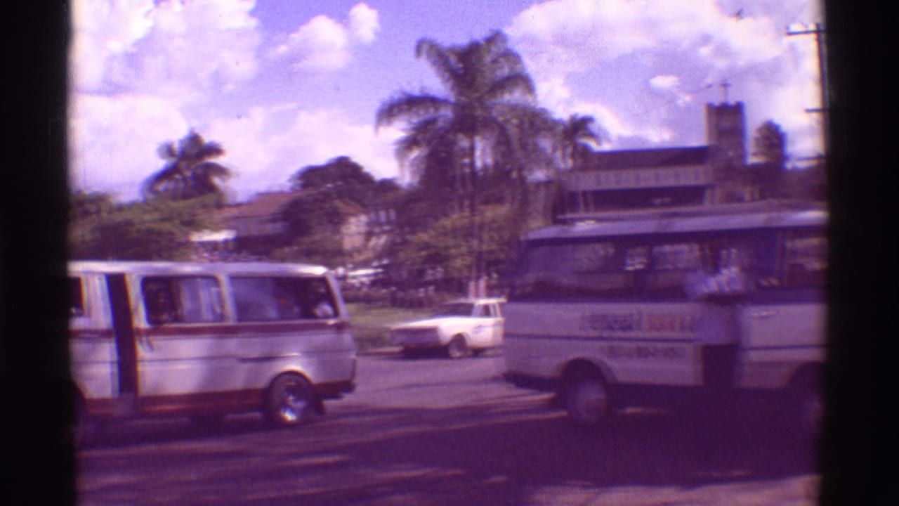 1971 Hawaii street view with tour vans, a white car, palm trees, utility poles, and a low building seen from a handheld 8mm camera.