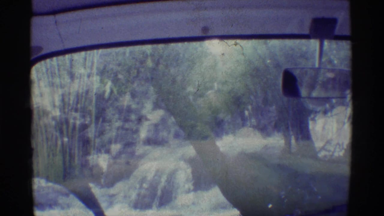 1971 Mauna Ulu, Hawaii home-movie frame from inside a car, showing a hand near the wheel, rearview mirror, windshield, and roadside trees.