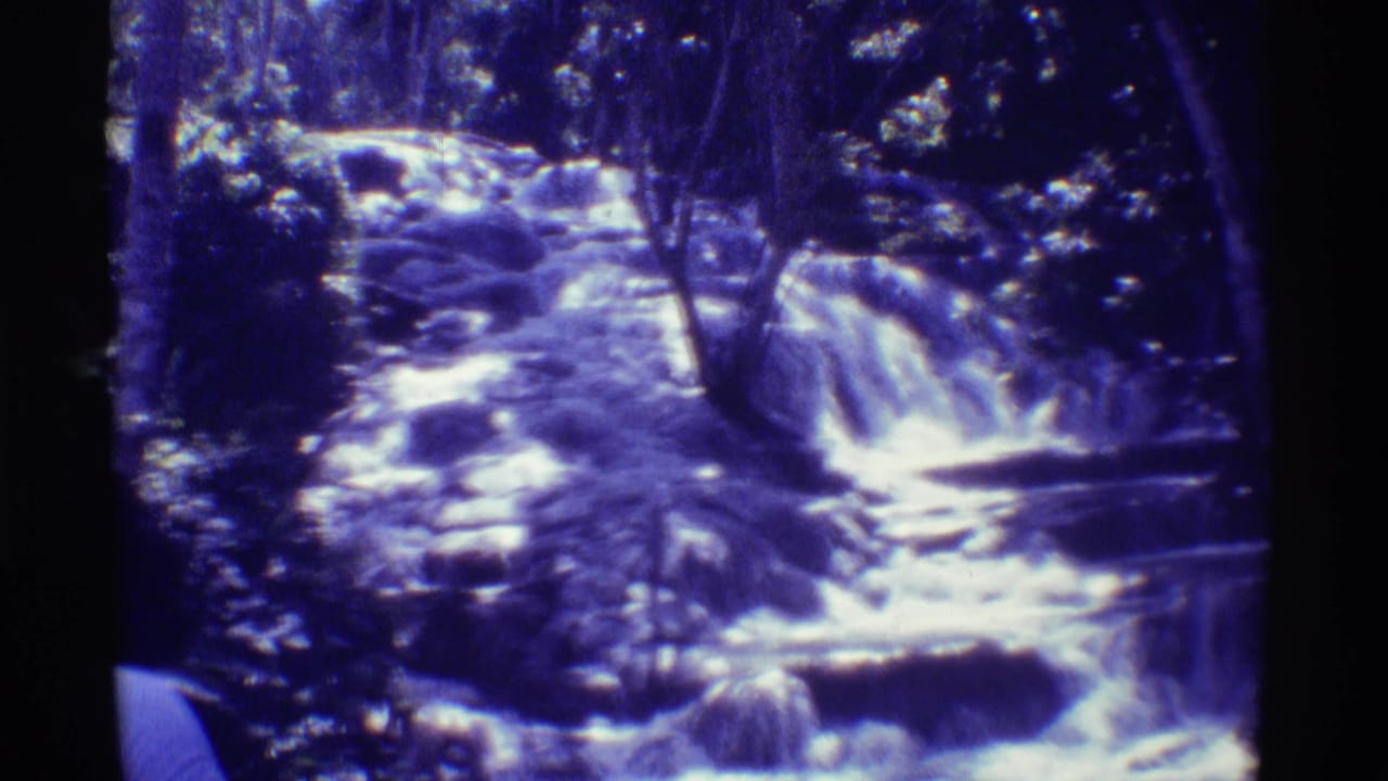 1971 Mauna Ulu, Hawaii home-movie frame of a low jungle waterfall flowing across rocks in blue-purple 8mm color.