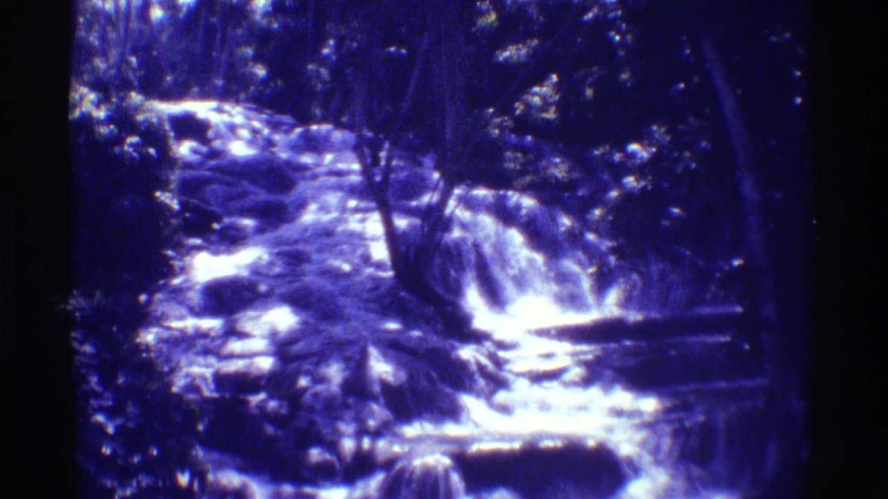 Blue-purple 8mm frame from Mauna Ulu showing a small waterfall and rocky stream surrounded by dense trees.