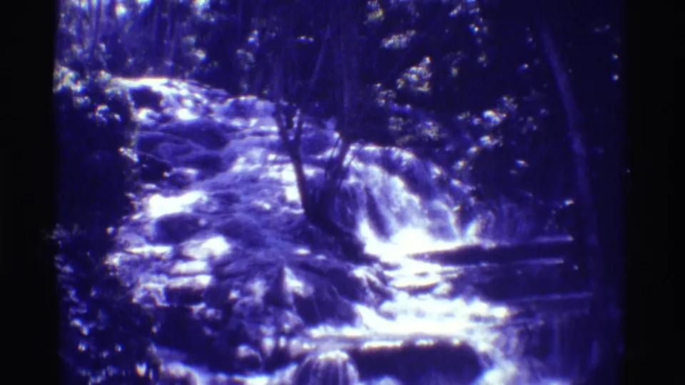 Blue-purple 8mm frame from Mauna Ulu showing a small waterfall and rocky stream surrounded by dense trees.