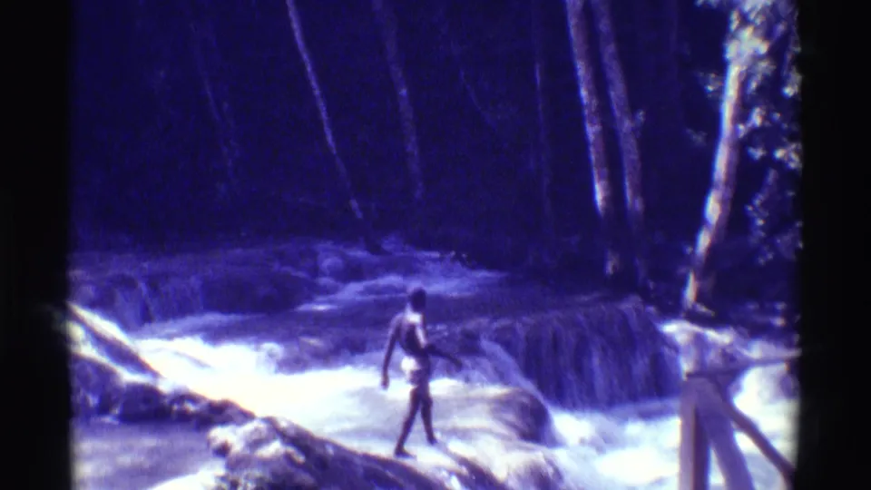 1971 Hawaii home-movie frame showing a person in yellow shorts stepping through a swift forest stream with rocks and trees around them.
