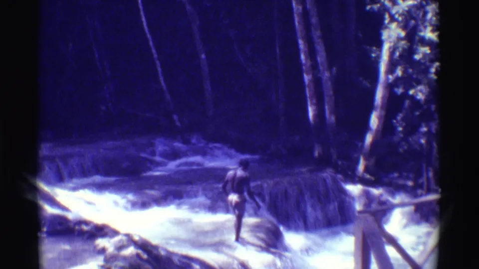 1971 Hawaii frame with a person moving across a small cascade in a wooded stream, filmed with a strong purple-blue cast.