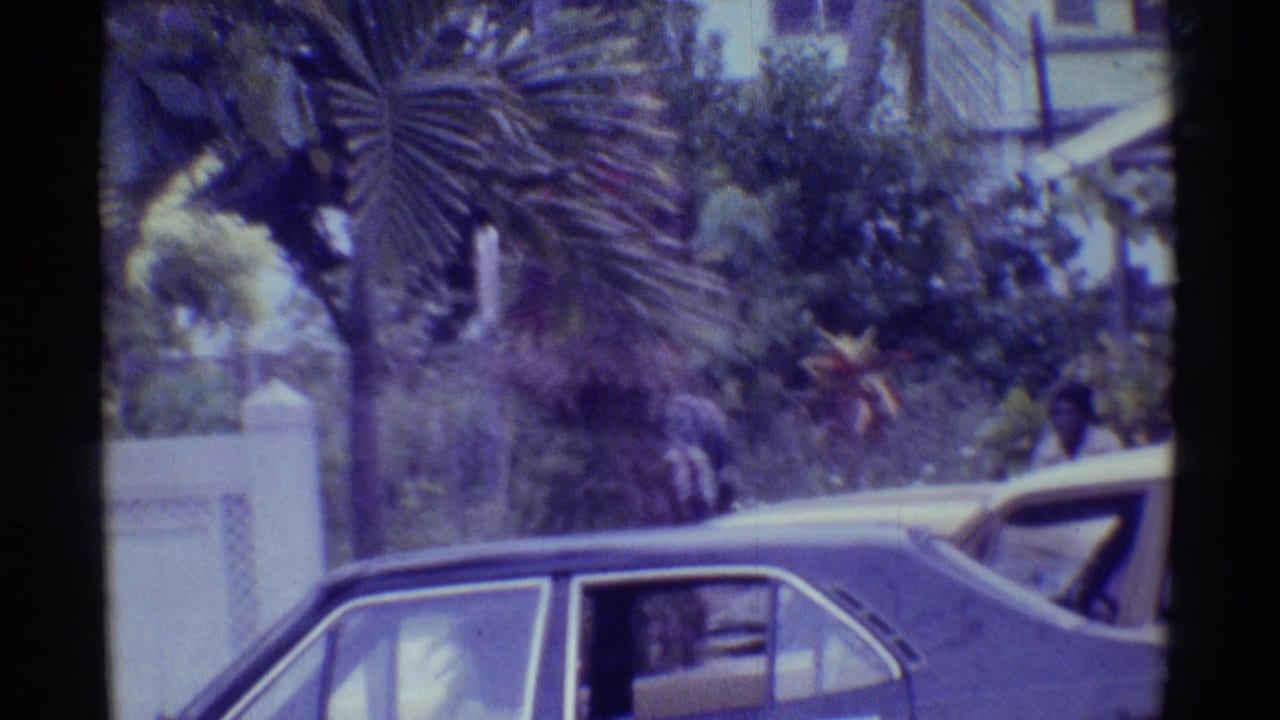 1971 Hawaii home-movie street view showing older cars beside a low wall, tropical planting, palm foliage, and a pedestrian in the distance.