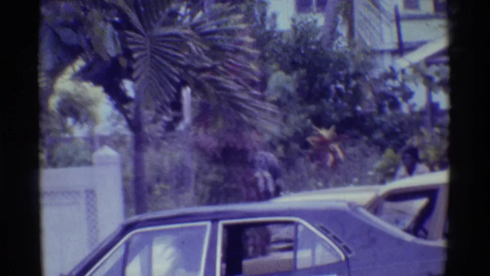 1971 Hawaii home-movie street view showing older cars beside a low wall, tropical planting, palm foliage, and a pedestrian in the distance.