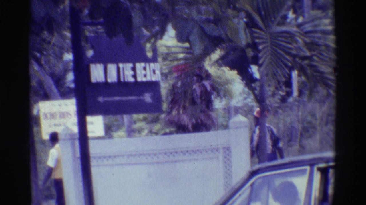 1971 Hawaii home-movie frame with a partially readable inn sign, pedestrians, a wall, palm trees, and an older car at the roadside.