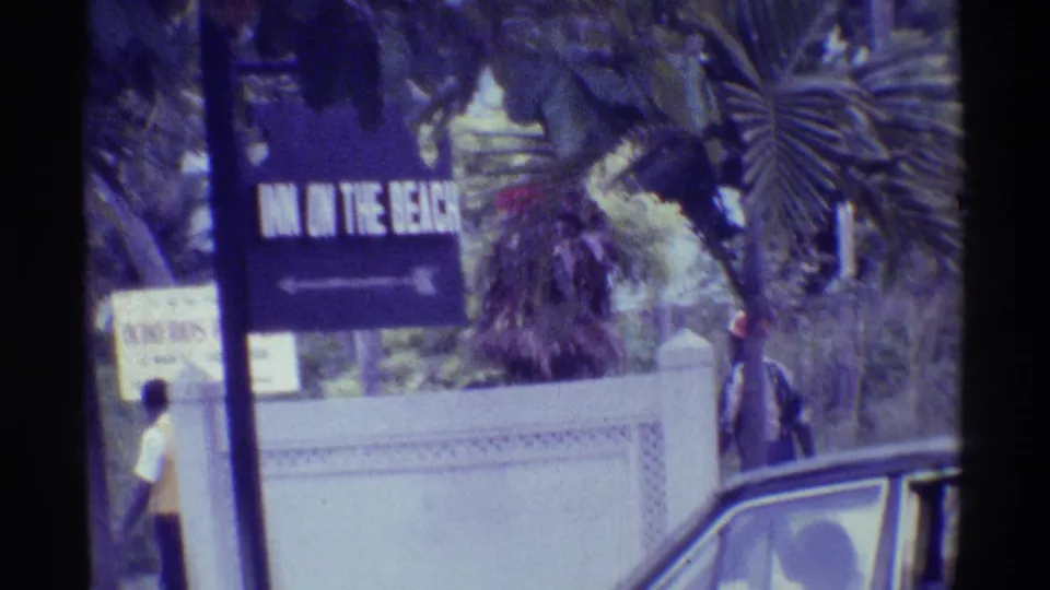 1971 Hawaii home-movie frame with a partially readable inn sign, pedestrians, a wall, palm trees, and an older car at the roadside.