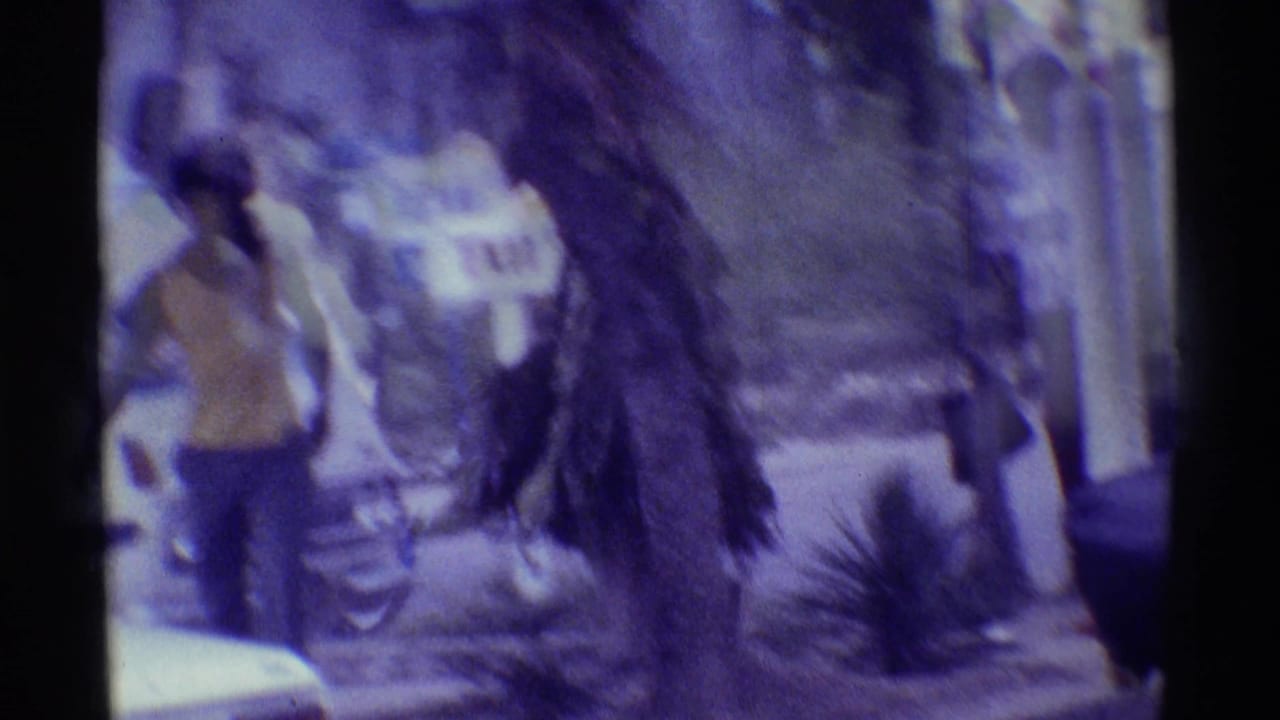 1971 Hawaii home-movie frame showing a man in a yellow top walking past tropical plants near a roadside inn sign.