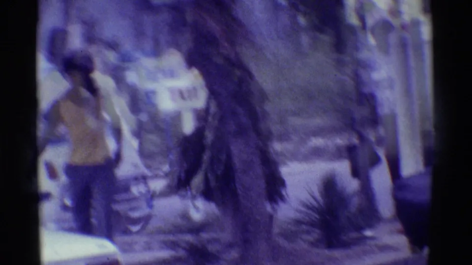 1971 Hawaii home-movie frame showing a man in a yellow top walking past tropical plants near a roadside inn sign.