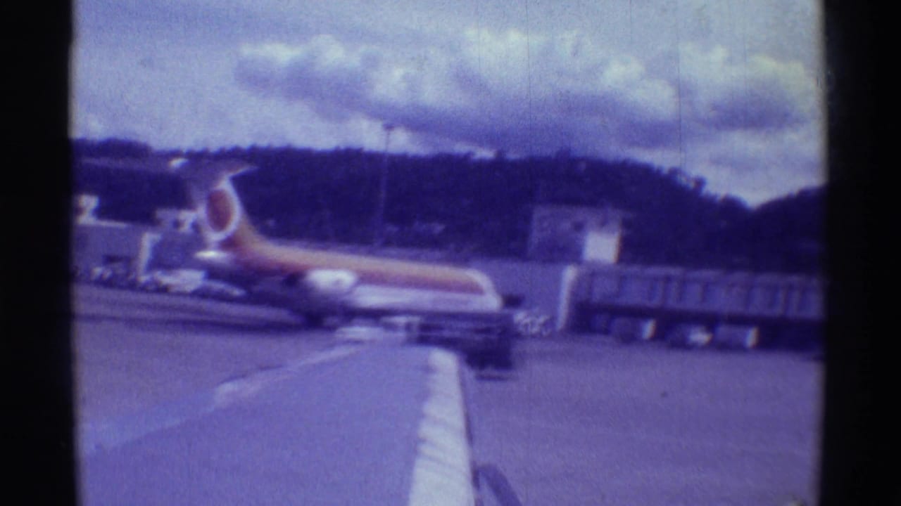 1971 Hawaii home-movie frame showing a small aircraft on tarmac with low hills, clouds, and airport buildings.