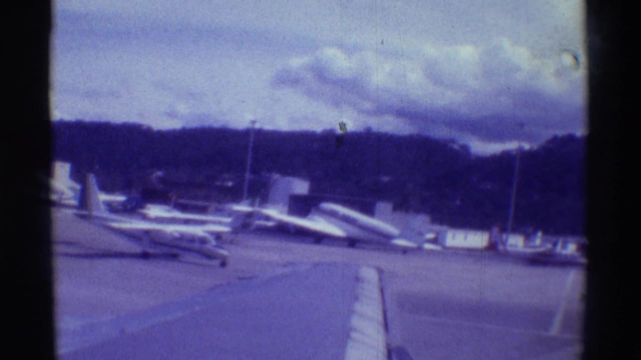 Small aircraft parked on a tarmac beneath a cloudy sky and wooded hillside.