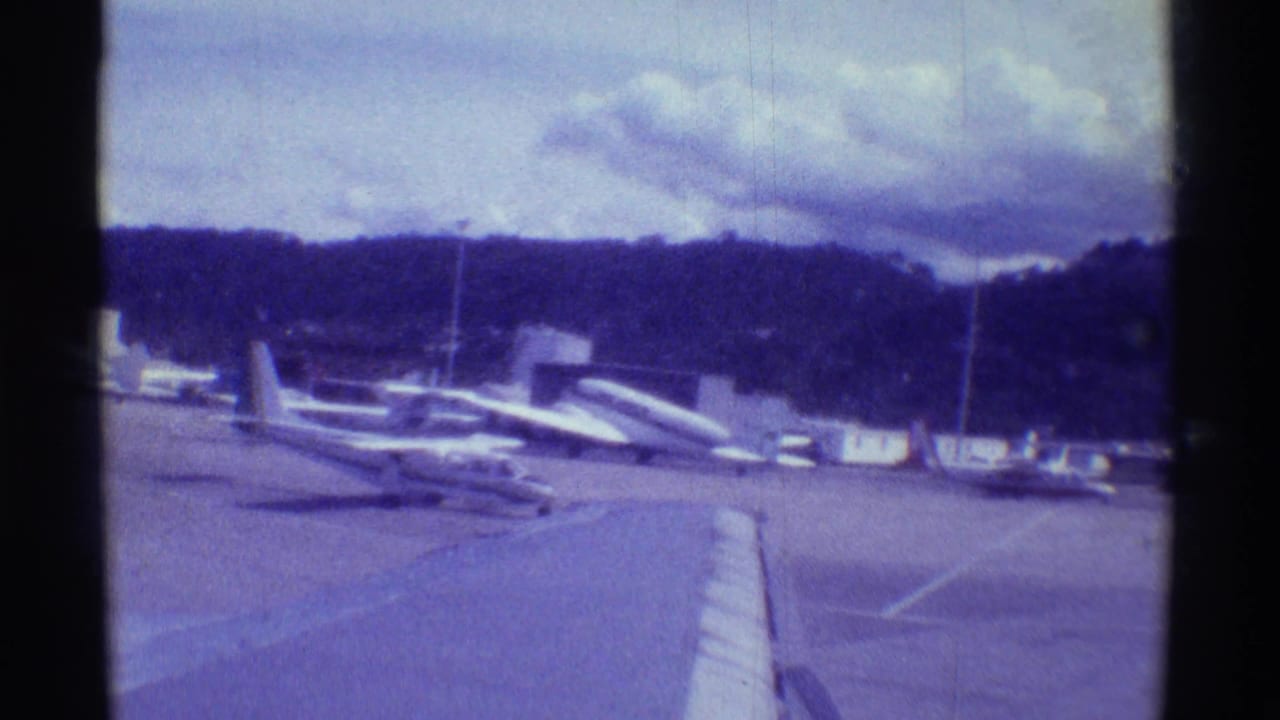 1971 Hawaii airport view with parked small planes, a larger aircraft shape, runway pavement, and hillside in faded 8mm color.
