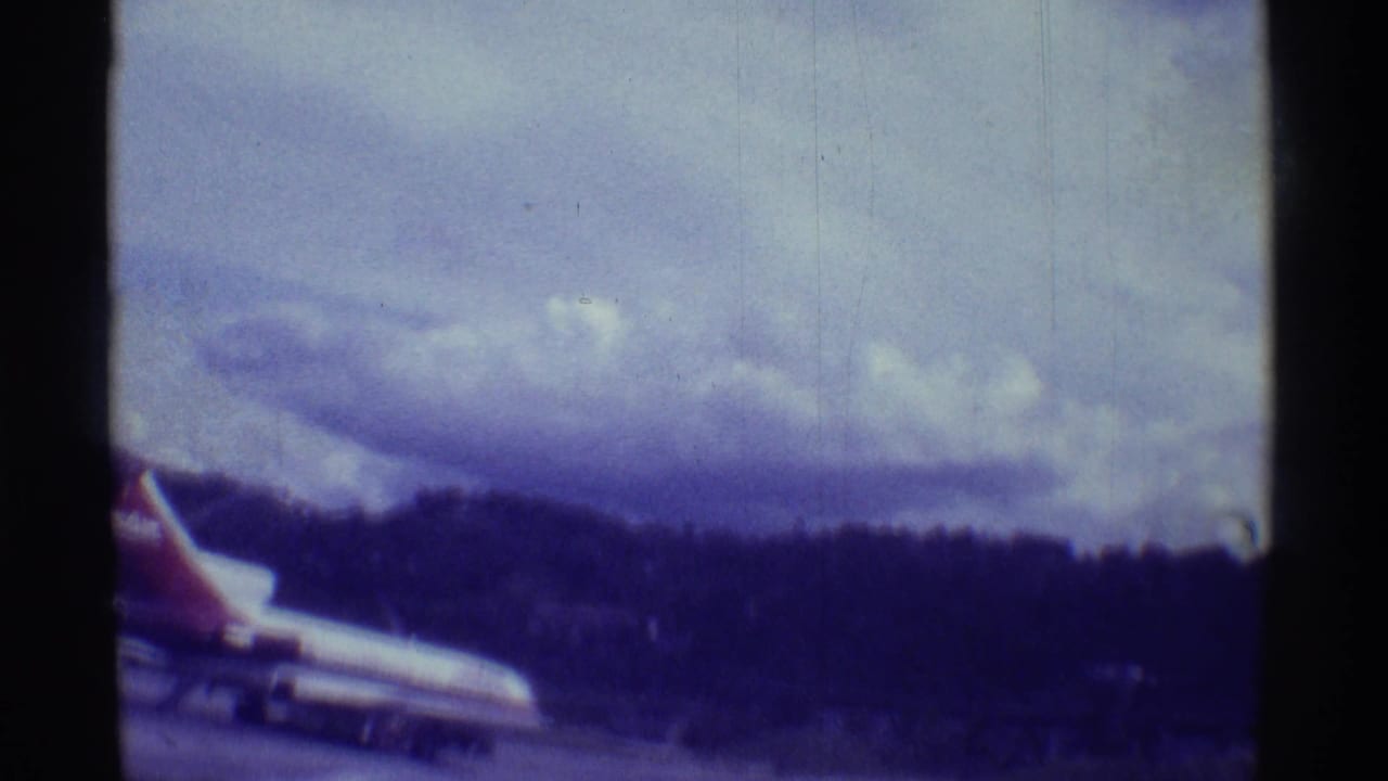 1971 Hawaii 8mm frame with a larger jet partly entering the frame, runway pavement, cloudy sky, and tree-covered hillside.