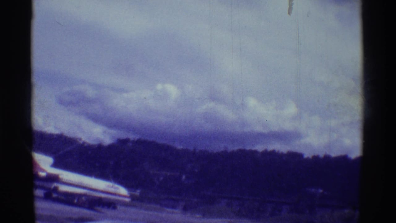 1971 Hawaii airport frame showing a larger aircraft near the runway, with a cloudy sky, hillside, and visible 8mm film wear.