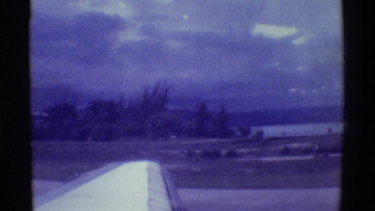 1971 8mm Hawaii takeoff view with a white airplane wing in the foreground and coastline under a cloudy sky.