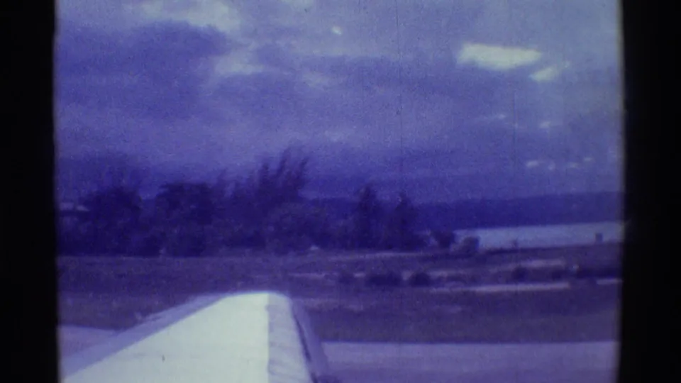 1971 8mm Hawaii takeoff view with a white airplane wing in the foreground and coastline under a cloudy sky.