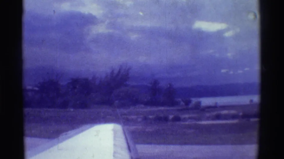Small aircraft takeoff view in Hawaii, with wing foreground, shoreline vegetation, ocean, and blue-purple 8mm color cast.