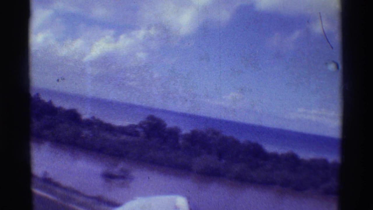 1971 8mm frame from a small plane over Hawaii, showing a tilted coastline, ocean, clouds, and wing edge.