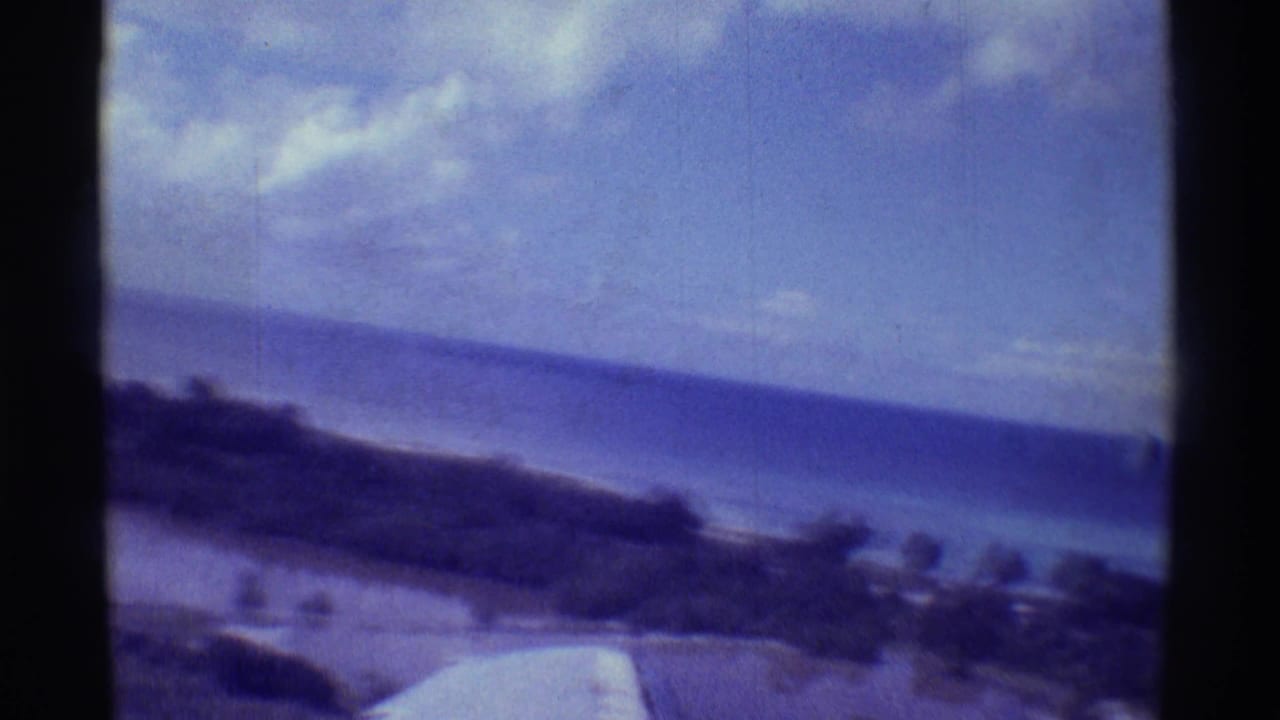 Small-plane view over the Hawaiian coast, with ocean, shoreline, wing foreground, and soft blue 8mm film color.