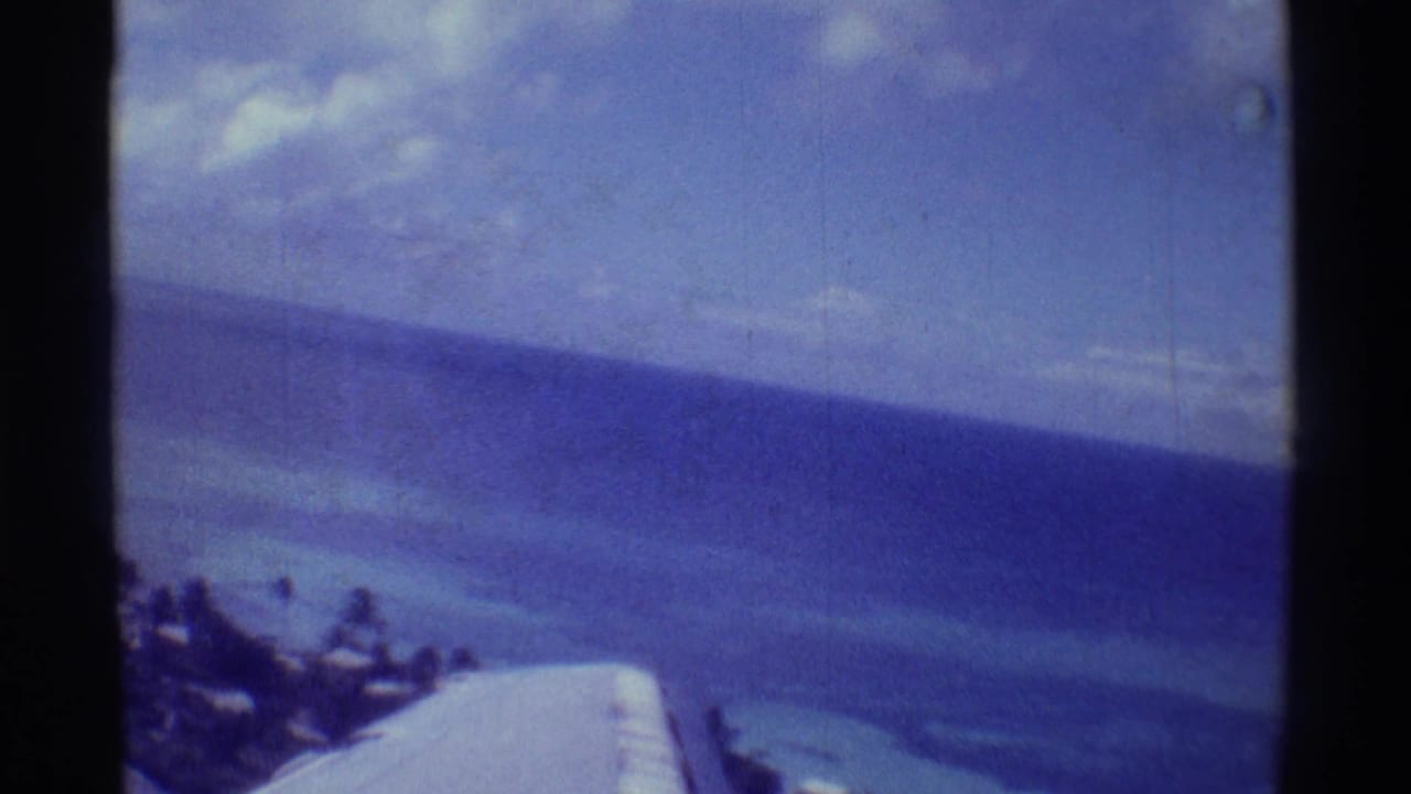 1971 Hawaii 8mm frame showing a soft aerial view of reef-toned water, shoreline trees, and plane wing.