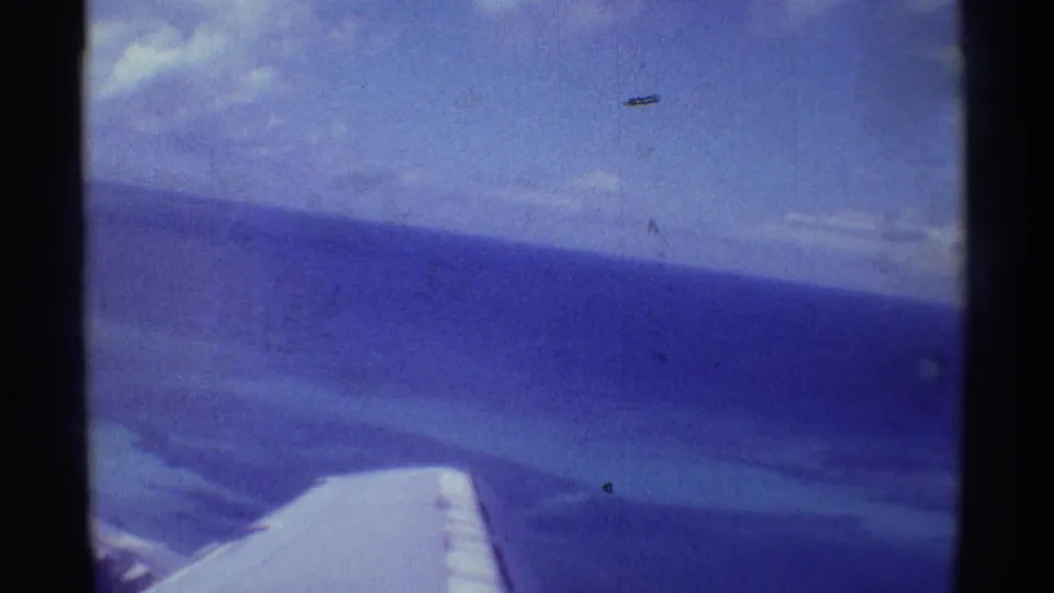 1971 8mm Hawaii travel frame showing an airplane wing through a window above blue ocean, clouds, and a distant horizon.