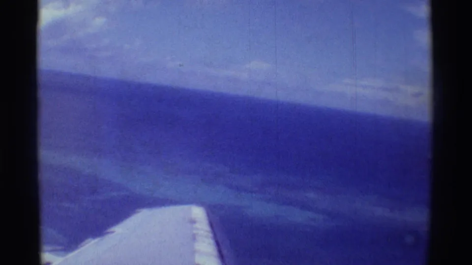 1971 8mm frame from an airplane window, with the wing low in frame and blue ocean stretching toward the horizon.